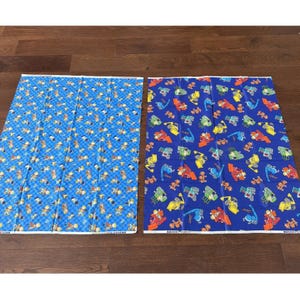 May include: Two rectangular pieces of fabric. One is blue with a checkered pattern and cartoon figures. The other is blue with a pattern of construction vehicles in various colors. Both have white borders.