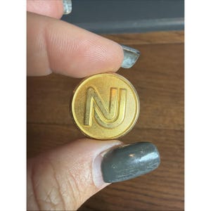 May include: A circular gold-toned button featuring a raised letter N in the center. The surface of the button has a fine, linear textured pattern radiating from the center, and the letter N has a smooth, polished finish with a raised border.