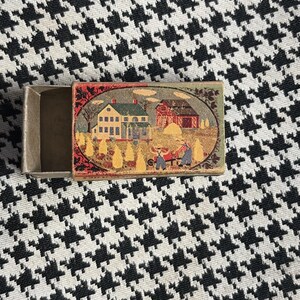 May include: An open matchbox with a vintage illustration of a farm scene. The artwork depicts a white house, a red barn, and figures working in a field. The box is set against a black and white houndstooth pattern.