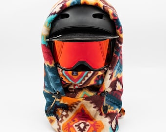 May include: A colorful fleece balaclava with a geometric pattern. The balaclava is worn over a black ski helmet with a pair of red ski goggles.