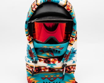 May include: A black ski helmet and red-lensed goggles are encased in a patterned fleece balaclava. The balaclava features a turquoise, white, orange, and red geometric design. Drawstrings are visible at the neck.