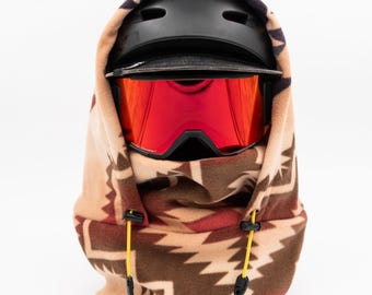 Könnte beinhalten: Ein brauner, beigefarbener und roter Fleece-Halswärmer mit geometrischem Muster. Der Halswärmer hat eine Kapuze und zwei Kordeln mit schwarzen Stoppern. Ein schwarzer Skihelm mit schwarzem Visier und roten Skibrillen wird über dem Halswärmer getragen.