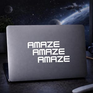 Könnte beinhalten: Ein grauer Laptop mit dem Wort "AMAZE" dreimal weiß übereinander gestapelt. Der Hintergrund zeigt eine Weltraumszene mit Planeten und einem Raumschiff.