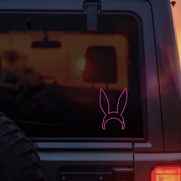 Könnte beinhalten: Ein leuchtend pinkfarbener Vinyl-Aufkleber, der den Umriss eines Paares langer Hasenohren an einem Stirnband zeigt. Der Aufkleber ist auf der Heckscheibe eines dunklen Fahrzeugs angebracht und zeigt ein klares, grafisches Design.
