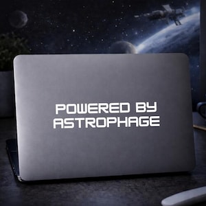 May include: A gray laptop with the text "POWERED BY ASTROPHAGE" in white. The laptop is open and sitting on a dark surface. The background shows a space scene with planets and a spaceship.