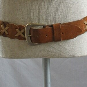 May include: A brown leather belt with a silver buckle and a braided design. The belt is made of multiple pieces of leather that are woven together.