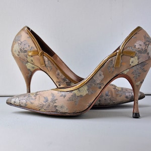 May include: A pair of high-heeled shoes with a floral pattern. The shoes are made of a light brown fabric with a gold trim. The heels are very high and thin.
