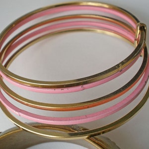 May include: A multi-layered bangle bracelet featuring thin, circular gold-tone metal bands alternating with light pink enamel-coated bands. The structure consists of several interconnected rings that form a single cohesive piece of jewelry.