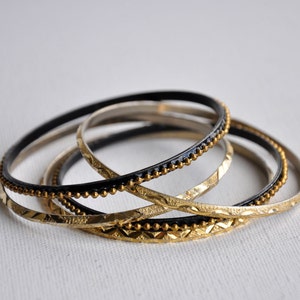 May include: A set of four stacked bangles featuring two black bands with a raised gold-tone beaded texture and two gold-tone bands with a textured, faceted surface. The bracelets are thin, circular, and designed to be worn together as a set.
