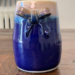 May include: A ceramic vase with a gradient of colors, transitioning from a light tan at the top to a deep blue at the base. A sculpted bow detail is affixed to the front. The vase is likely handmade and has a glossy finish.