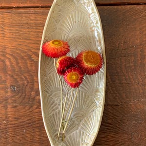 May include: A cream-colored, oval-shaped ceramic dish with a textured fan design. The dish holds a small bouquet of dried flowers with red and orange blooms. The arrangement sits on a dark wooden surface.