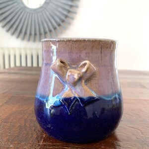 May include: A handmade ceramic vase with a bow design. The vase features a gradient of colors, transitioning from a dark blue base to a purple midsection and a brown rim. The bow is a lighter brown color.