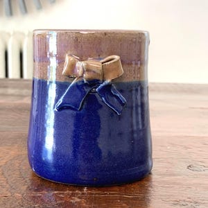 May include: A handmade ceramic vase with a two-tone design. The bottom half is a deep blue, transitioning to a brown-purple at the top. A decorative bow is affixed to the front. The vase is likely for home decor.