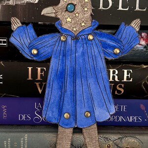 May include: A whimsical paper cutout of a bird-like figure wearing a blue cape with gold buttons. The bird's head is adorned with stars and a large blue eye. The figure is standing in front of a stack of books.