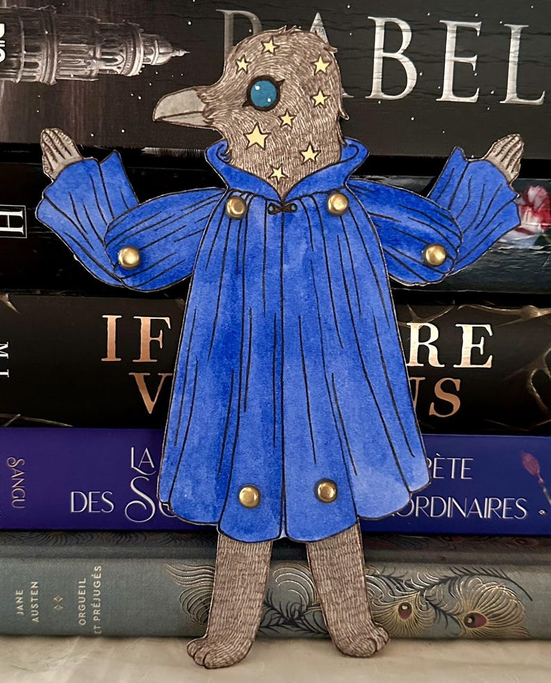 May include: A whimsical paper cutout of a bird-like figure wearing a blue cape with gold buttons. The bird's head is adorned with stars and a large blue eye. The figure is standing in front of a stack of books.
