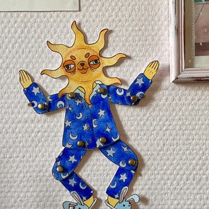 May include: A whimsical wall decoration featuring a sun-faced figure in blue pajamas adorned with moons and stars. The figure has outstretched arms and is wearing bunny slippers. The artwork is attached to the wall with brass fasteners.