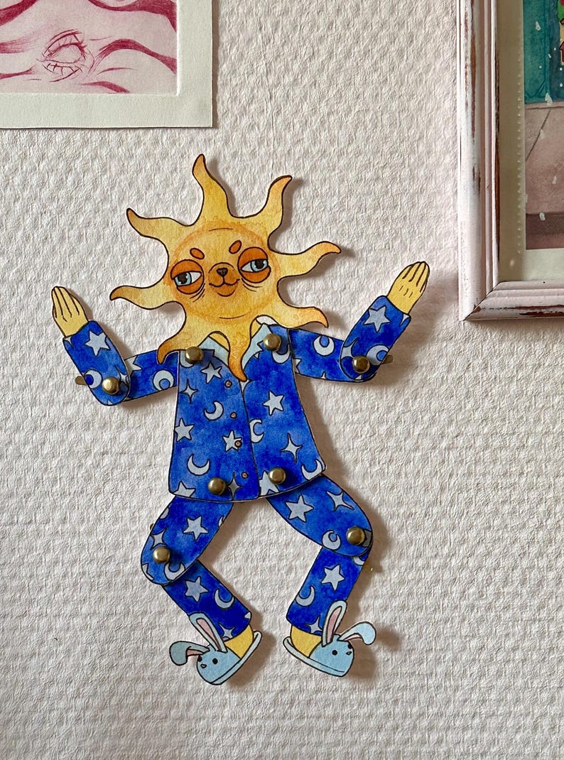 May include: A whimsical wall decoration featuring a sun-faced figure in blue pajamas adorned with moons and stars. The figure has outstretched arms and is wearing bunny slippers. The artwork is attached to the wall with brass fasteners.