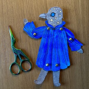 May include: A whimsical paper cutout of a bird-like figure in a blue cape with gold buttons, and a pair of green and iridescent scissors. The bird has a gray head with star accents and a blue eye. The items are on a wooden surface.