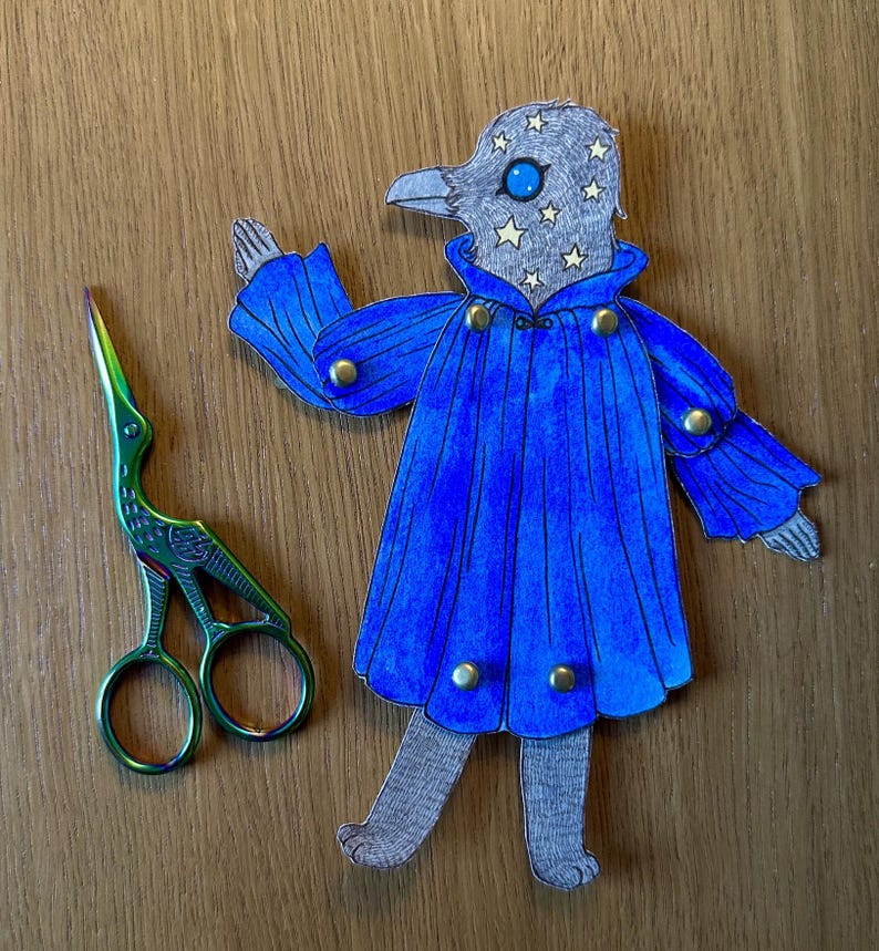 May include: A whimsical paper cutout of a bird-like figure in a blue cape with gold buttons, and a pair of green and iridescent scissors. The bird has a gray head with star accents and a blue eye. The items are on a wooden surface.