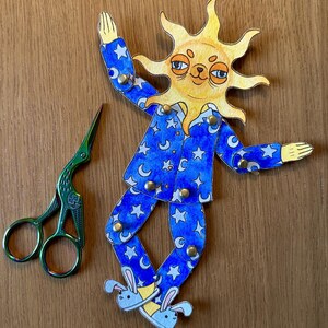 May include: A whimsical paper cutout of a sun wearing blue pajamas with white stars and crescent moons. The sun has a smiling face and is wearing bunny slippers. A pair of green scissors is next to the sun cutout.