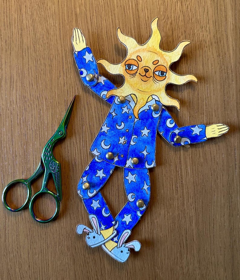 May include: A whimsical paper cutout of a sun wearing blue pajamas with white stars and crescent moons. The sun has a smiling face and is wearing bunny slippers. A pair of green scissors is next to the sun cutout.