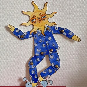 May include: A whimsical, hand-painted paper doll of a sun wearing blue pajamas with a crescent moon and star pattern. The sun has a smiling face and is wearing bunny slippers. The doll is jointed with brass fasteners.