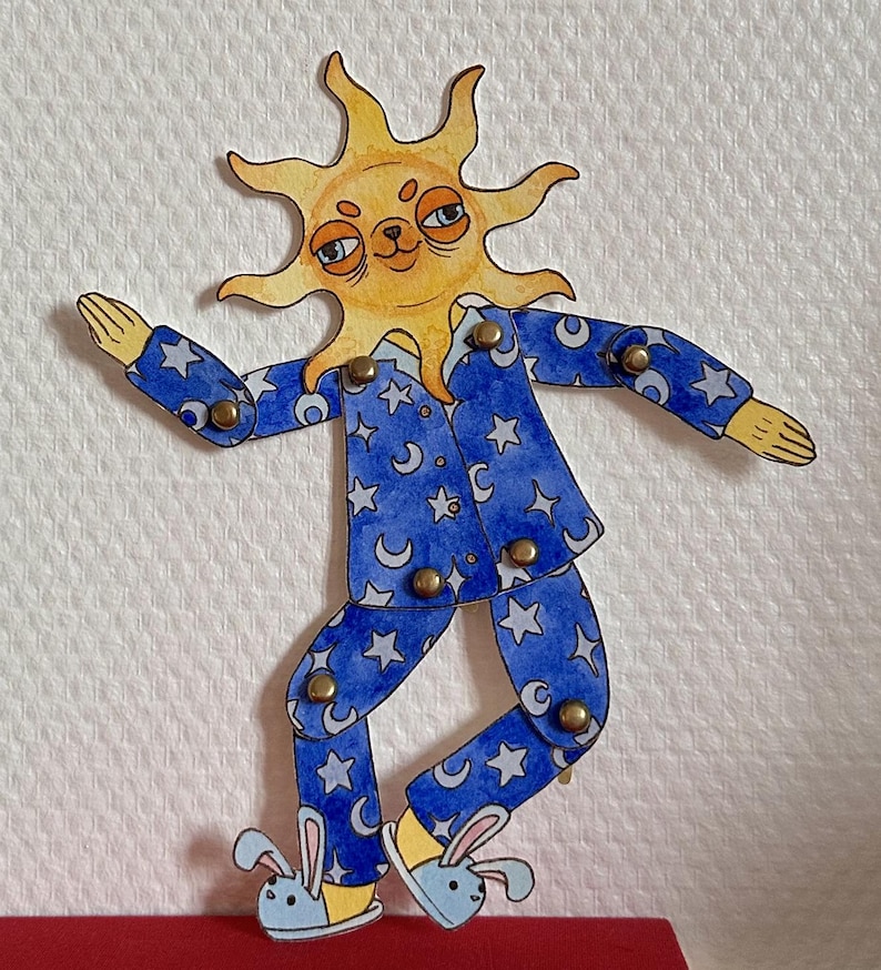 May include: A whimsical, hand-painted paper doll of a sun wearing blue pajamas with a crescent moon and star pattern. The sun has a smiling face and is wearing bunny slippers. The doll is jointed with brass fasteners.