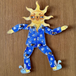 May include: A whimsical, articulated paper doll in the shape of a sun wearing blue pajamas. The pajamas are decorated with white stars and crescent moons. The doll's feet are bunny slippers. The sun has a smiling face.