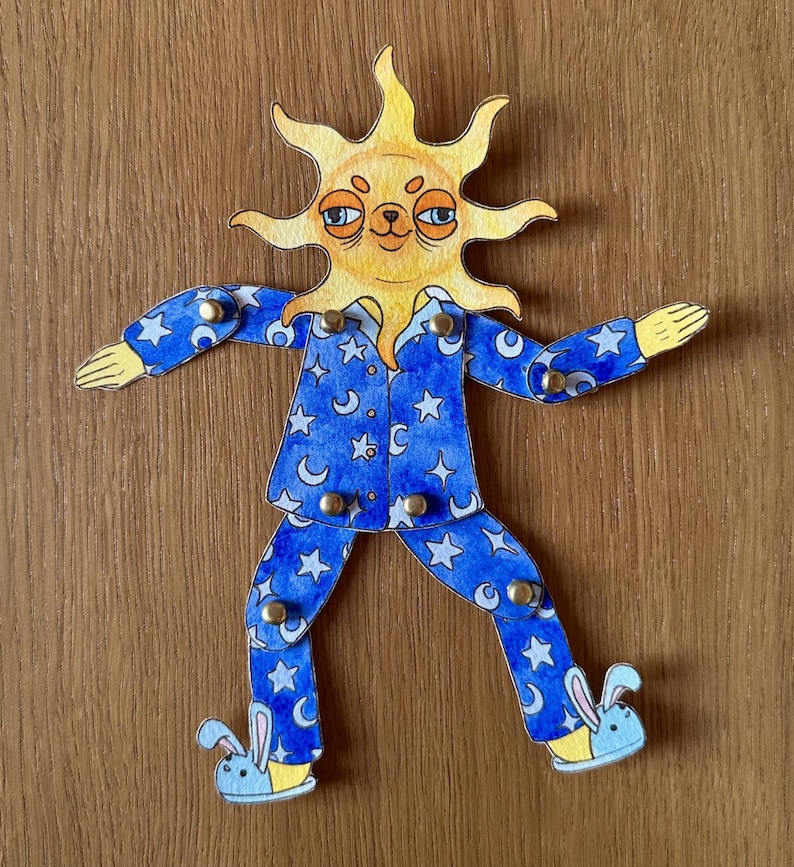 May include: A whimsical, articulated paper doll in the shape of a sun wearing blue pajamas. The pajamas are decorated with white stars and crescent moons. The doll's feet are bunny slippers. The sun has a smiling face.