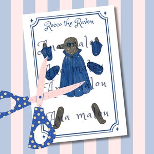 May include: A paper cutout featuring a watercolor illustration of "Rocco the Raven" with a blue cape and accessories. The image is on a white background with a blue border, and is cut with a pair of blue and yellow polka dot scissors.