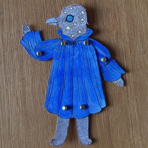 May include: A whimsical paper cutout of a bird-like figure with a blue cape and star-studded head. The figure has a gray head with a blue eye and a beak. The blue cape is adorned with gold-colored buttons. The figure is standing on a wooden surface.
