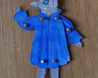 Puede incluir: Una figura de papel de un pájaro con una capa azul y una cabeza salpicada de estrellas. La figura tiene una cabeza gris con un ojo azul y un pico. La capa azul está adornada con botones dorados. La figura está sobre una superficie de madera.