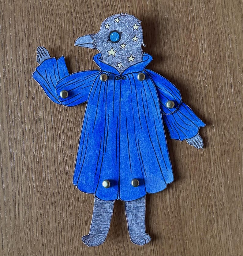 May include: A whimsical paper cutout of a bird-like figure with a blue cape and star-studded head. The figure has a gray head with a blue eye and a beak. The blue cape is adorned with gold-colored buttons. The figure is standing on a wooden surface.