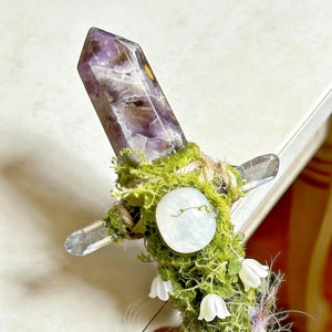 May include: A decorative wand featuring a pointed amethyst crystal at the top, wrapped in green moss and twine. The handle is adorned with a smooth, round, pale white stone and small, white, bell-shaped flower accents attached to the mossy surface.