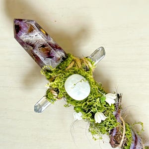 May include: A decorative wand featuring a large, pointed amethyst crystal at the top, accented with green moss, a smooth white stone, and small white flower blossoms. The handle is wrapped in natural twine and additional moss, creating a textured look.