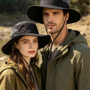 Pode incluir: Duas pessoas usando chapéus de sol pretos com abas largas e barbichos. Eles também estão usando jaquetas com capuz verde-oliva. Os chapéus têm um painel de malha para ventilação. O fundo é uma cena externa desfocada com árvores e água.