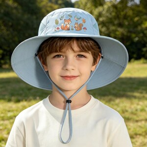 Puede incluir: Un sombrero de sol azul claro de ala ancha con un estampado gráfico de un zorro, una ardilla, un pájaro azul, hojas y setas en la parte frontal. El sombrero incluye una correa ajustable para la barbilla con un cierre negro de ajuste.