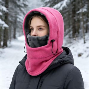 May include: A bright pink fleece balaclava featuring an integrated hood and a dark gray inner neck gaiter. The hood includes black adjustable drawstrings with toggles, providing full coverage for the head, neck, and face in cold weather.