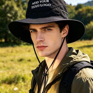 Può includere: Cappello da sole nero con tesa larga e sottogola. Il cappello ha la scritta bianca "ADD YOUR TEXT/LOGO". La persona indossa una giacca verde ed è all'aperto.