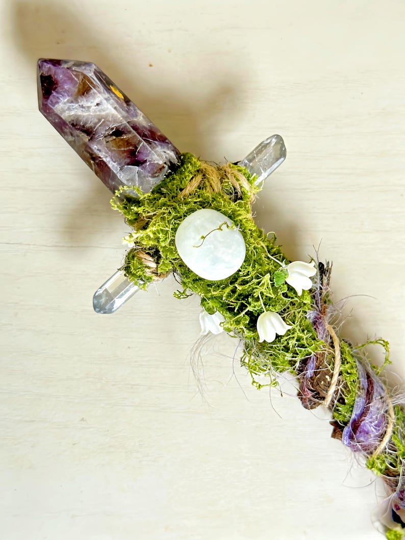 May include: A decorative wand featuring a large, pointed amethyst crystal at the top, accented with green moss, a smooth white stone, and small white flower blossoms. The handle is wrapped in natural twine and additional moss, creating a textured look.
