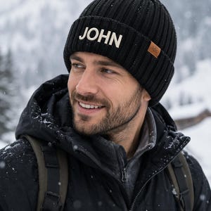 May include: A black ribbed knit beanie featuring the name JOHN in white capital letters on the front cuff. The beanie includes a small brown rectangular patch on the side with a mountain graphic logo, crafted from a thick, textured wool-like material.