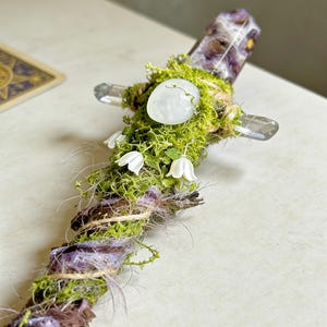 May include: A decorative wand featuring a central purple amethyst crystal point, accented with green moss, a smooth white stone, and small white bell-shaped flowers. The handle is wrapped in brown twine and purple fibers, creating a textured, organic appearance.