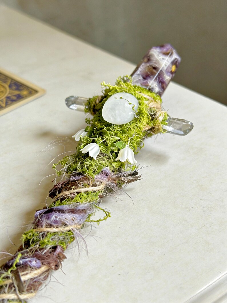 May include: A decorative wand featuring a central purple amethyst crystal point, accented with green moss, a smooth white stone, and small white bell-shaped flowers. The handle is wrapped in brown twine and purple fibers, creating a textured, organic appearance.