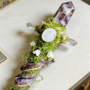 May include: A decorative wand featuring a pointed amethyst crystal at the top, a cross-guard made of clear quartz, and a handle wrapped in green moss, twine, and purple fibers. Small white bell-shaped flowers are attached to the mossy handle.