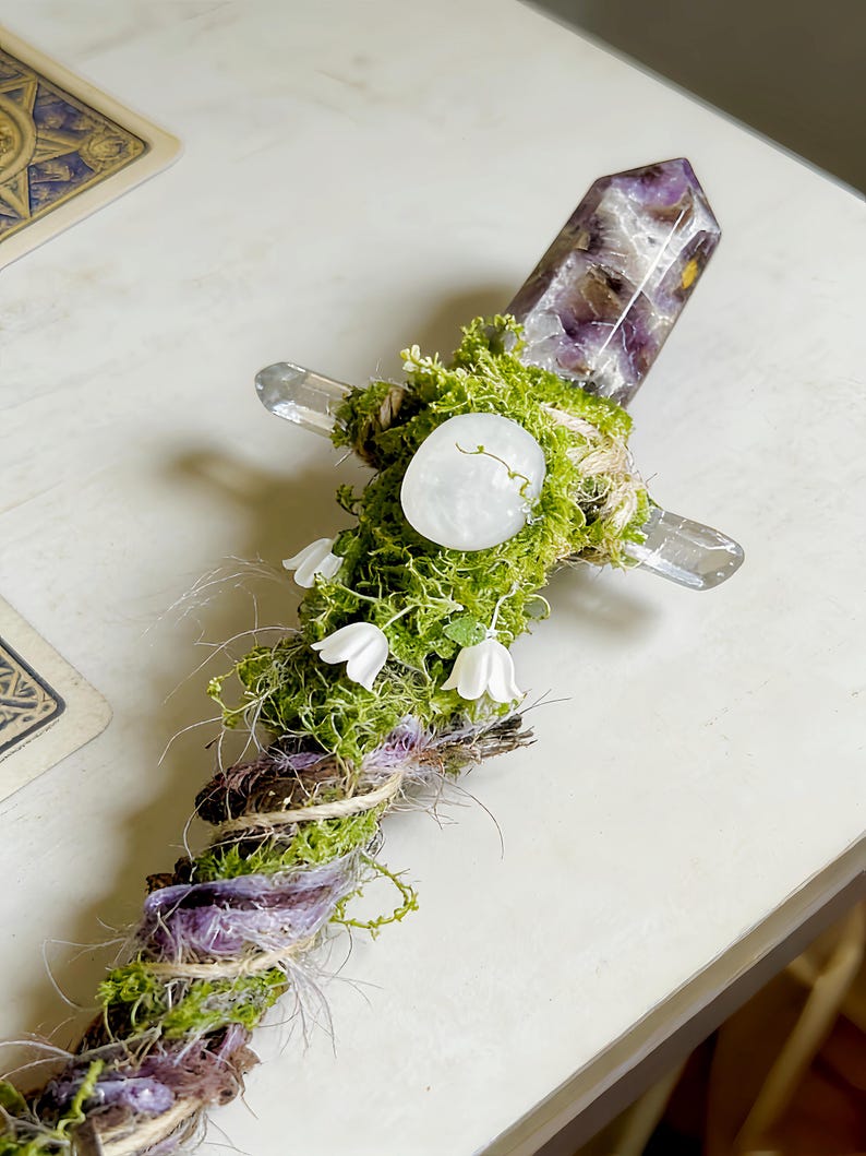 May include: A decorative wand featuring a pointed amethyst crystal at the top, a cross-guard made of clear quartz, and a handle wrapped in green moss, twine, and purple fibers. Small white bell-shaped flowers are attached to the mossy handle.