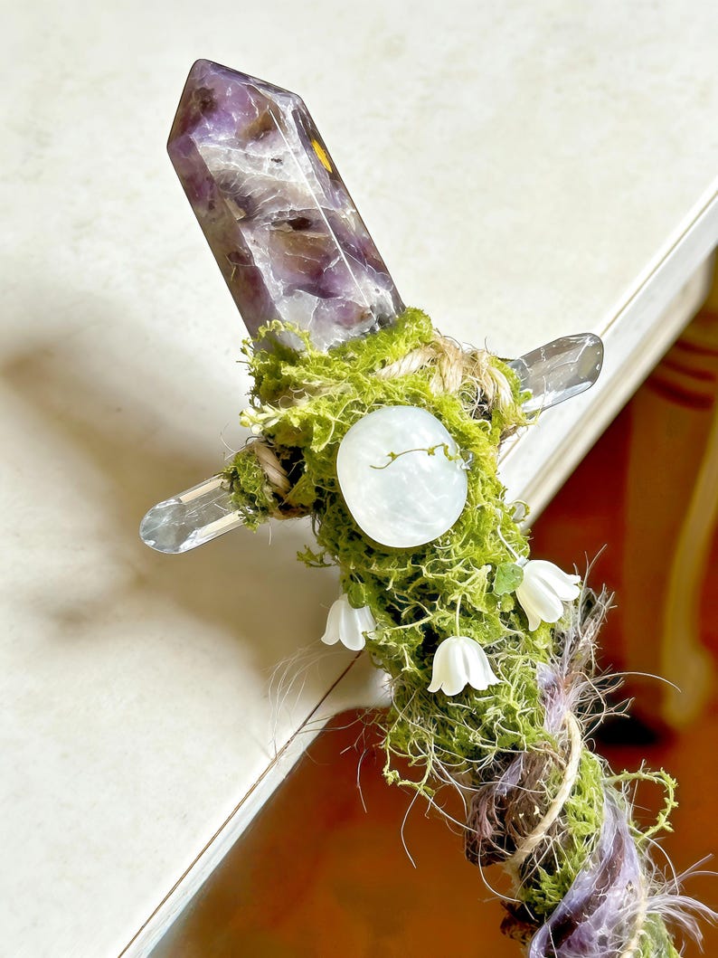 May include: A decorative wand featuring a pointed amethyst crystal at the top, wrapped in green moss and twine. The handle is adorned with a smooth, round, pale white stone and small, white, bell-shaped flower accents attached to the mossy surface.