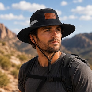 Peut inclure: Un chapeau de soleil noir à larges bords doté d'un écusson rectangulaire en cuir marron sur le devant avec le nom GRAYSON gravé en lettres noires. Le chapeau comprend une mentonnière noire réglable et une bande réfléchissante argentée.