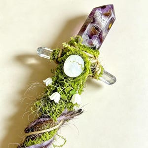 Peut inclure: Une baguette magique en cristal faite à la main avec une pointe d'améthyste violette, de la mousse verte, une pierre ronde blanche et de petites fleurs blanches en forme de cloche. Le manche est entouré de ficelle et d'un cordon violet.