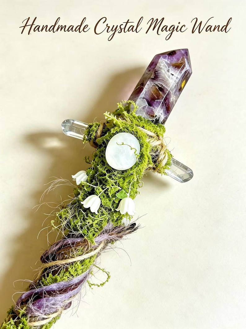 May include: A handmade crystal magic wand featuring a purple amethyst point, green moss, a white circular stone, and small white bell-shaped flowers. The handle is wrapped in twine and purple cord, with a clear crystal cross-guard accent.