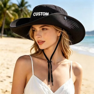 以下が含まれることがあります： 前面に白い大文字で「CUSTOM」とプリントされた、黒い広つばのサンハット。調節可能なトグル付きの黒いあご紐と、つばのサイドにスナップボタンが付いており、機能的なデザインとなっています。